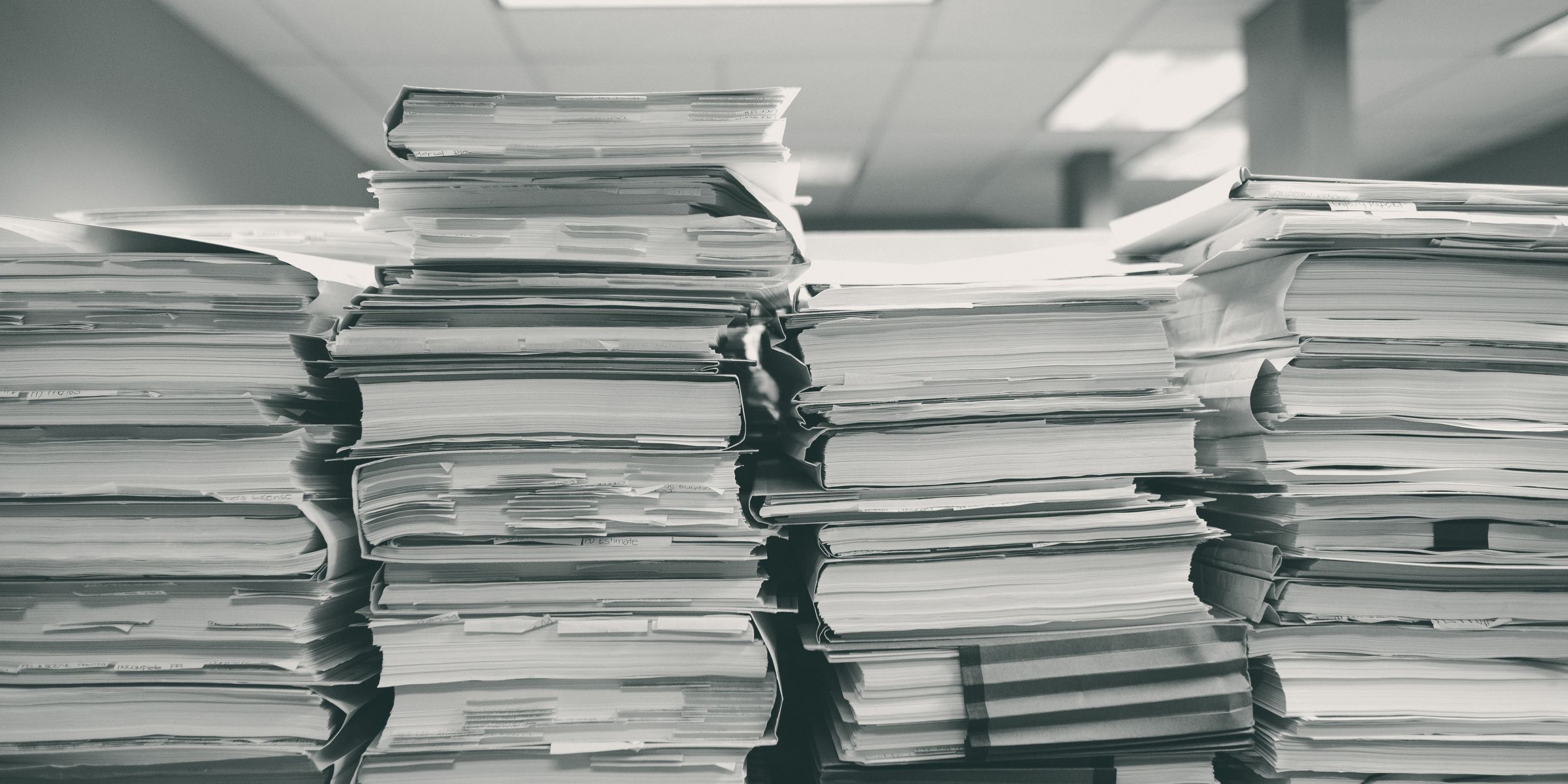 Pilhas altas de processos em papel, com etiquetas e marcadores, sobre uma mesa de escritório